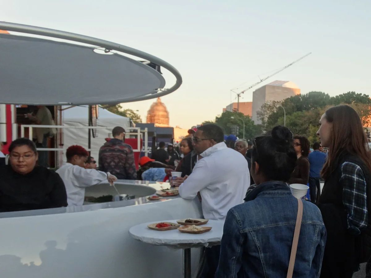 A PODULAR pod serving guests at an outdoor event, with staff behind the curved counter handing plates of food to a queue of customers